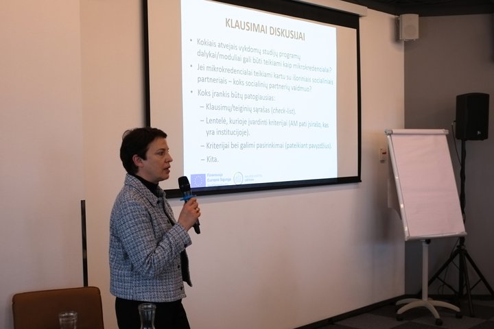 LT: Pranešėja Nora Skaburskienė laiko mikrofoną ir pristato temą, rodydama informaciją projektoriuje. EN: A presenter Nora Skaburskienė holds a microphone and shares information shown on the projected slide.