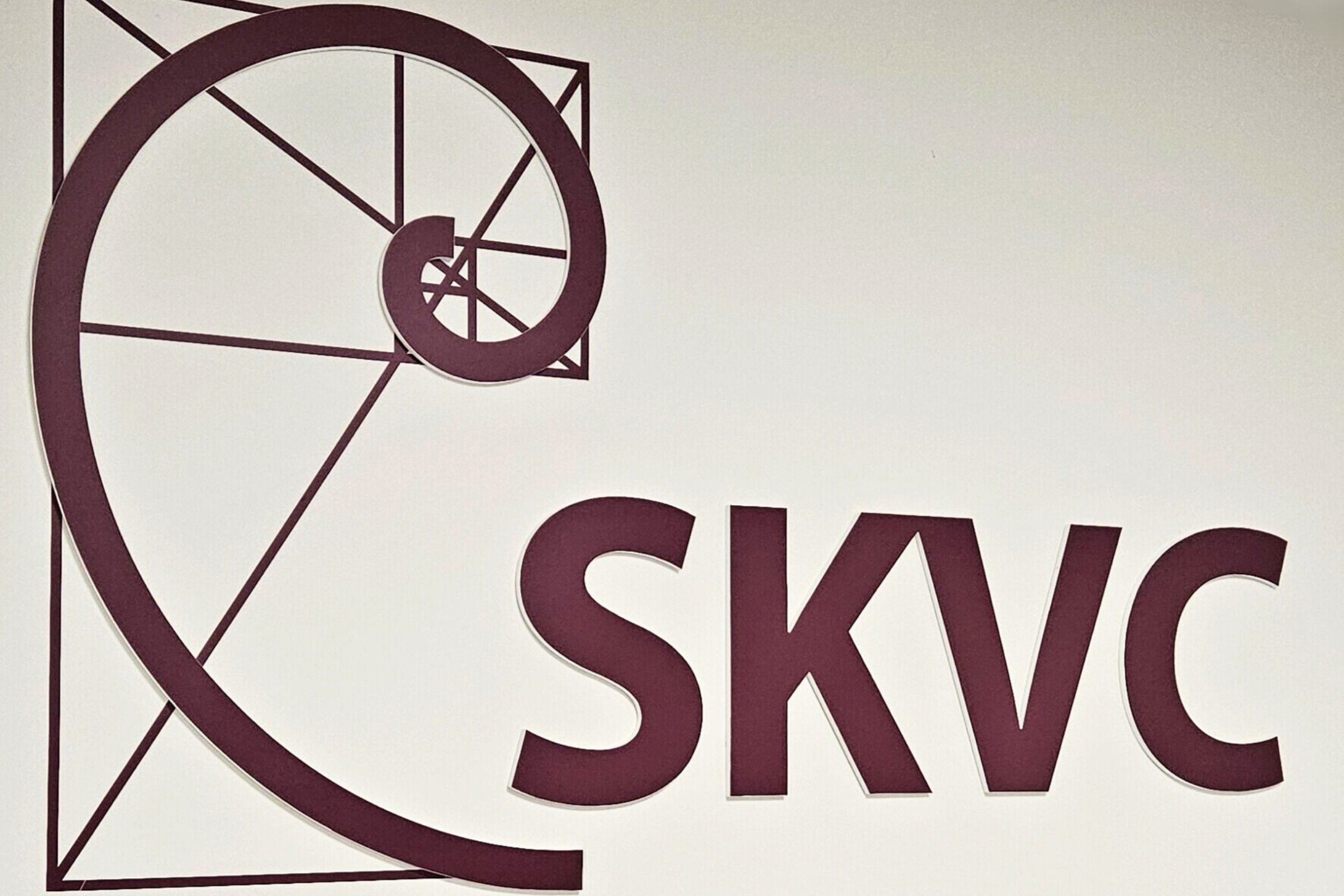 SKVC logotipas su stilizuota spirale ir raidėmis SKVC tamsiai vyšninės spalvos.