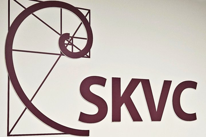 SKVC logotipas su stilizuota spirale ir raidėmis SKVC tamsiai vyšninės spalvos.