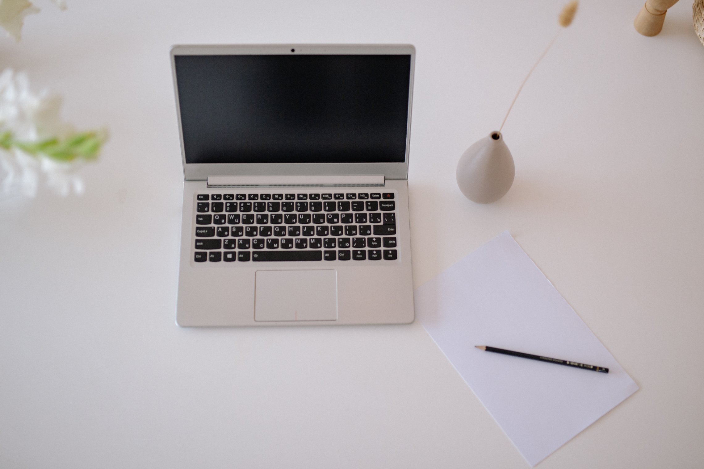 Lietuviškai: Švarus baltas stalas su nešiojamuoju kompiuteriu, popieriaus lapu su pieštuku ir vaza. English: A clean white desk featuring a laptop, a sheet of paper with a pencil, and a single vase.