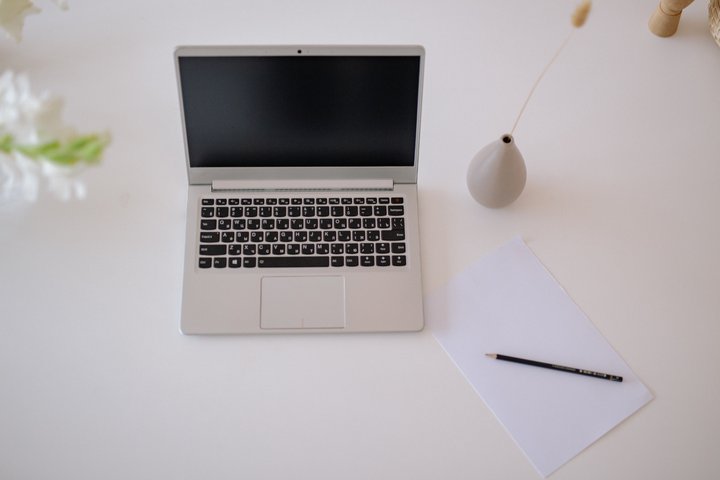 Lietuviškai: Švarus baltas stalas su nešiojamuoju kompiuteriu, popieriaus lapu su pieštuku ir vaza. English: A clean white desk featuring a laptop, a sheet of paper with a pencil, and a single vase.