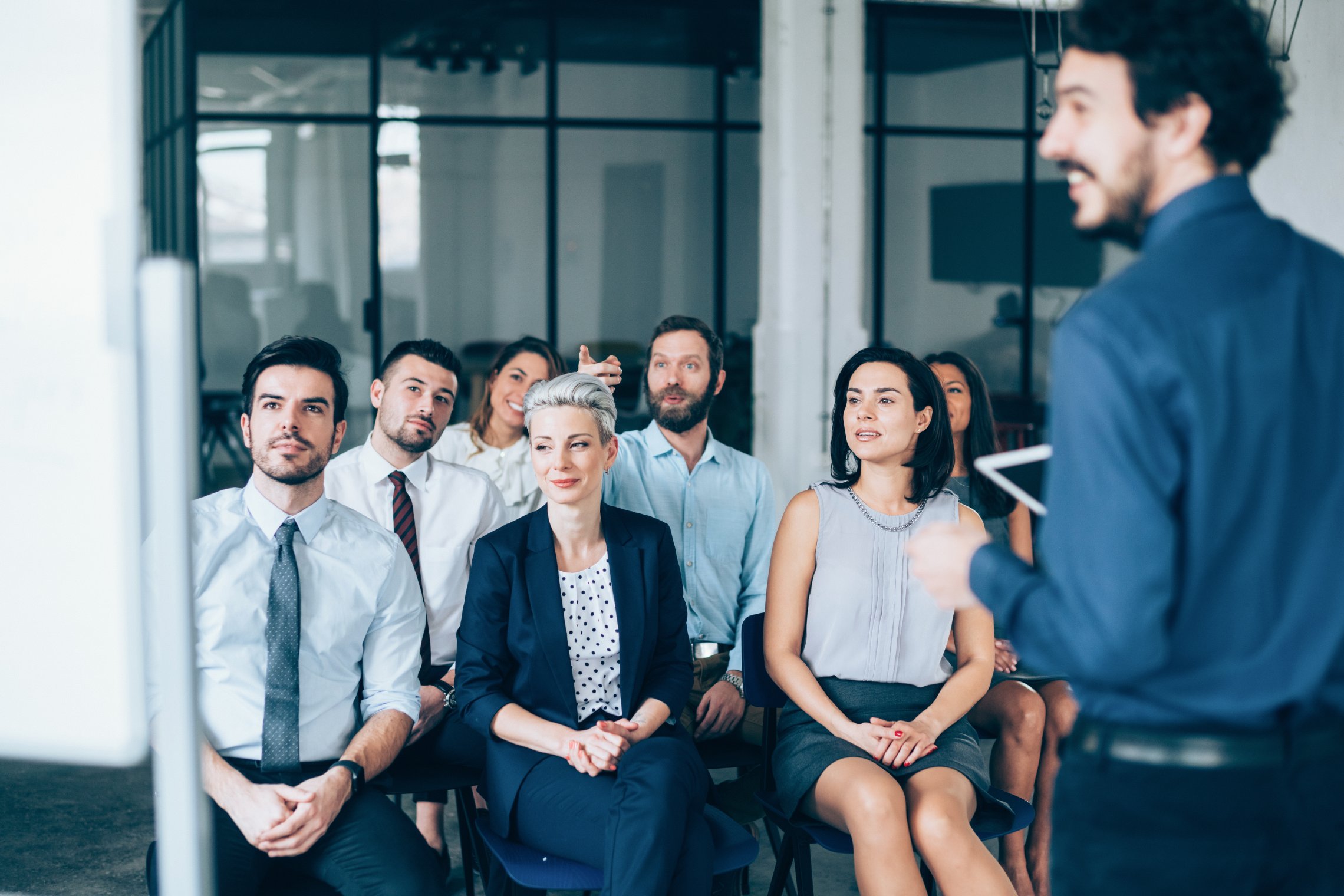 EN: An audience watching a presenter talk in a bright office setting
