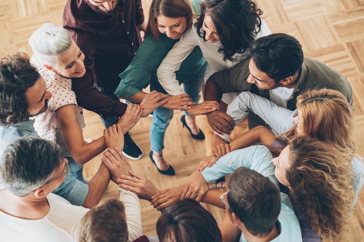 Lietuviškai: Žmonių grupė stovi ratu ir sudeda rankas į centrą, rodydami bendrystę ir komandą. English: A group of people standing in a circle with their hands joined in the center, showing unity and teamwork.