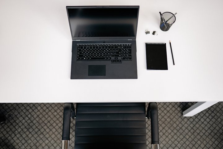 LT: Baltas darbo stalas iš viršaus su nešiojamuoju kompiuteriu, užrašine ir raštinės reikmenimis. / EN: Top view of a white desk with a laptop, notepad, and office supplies.