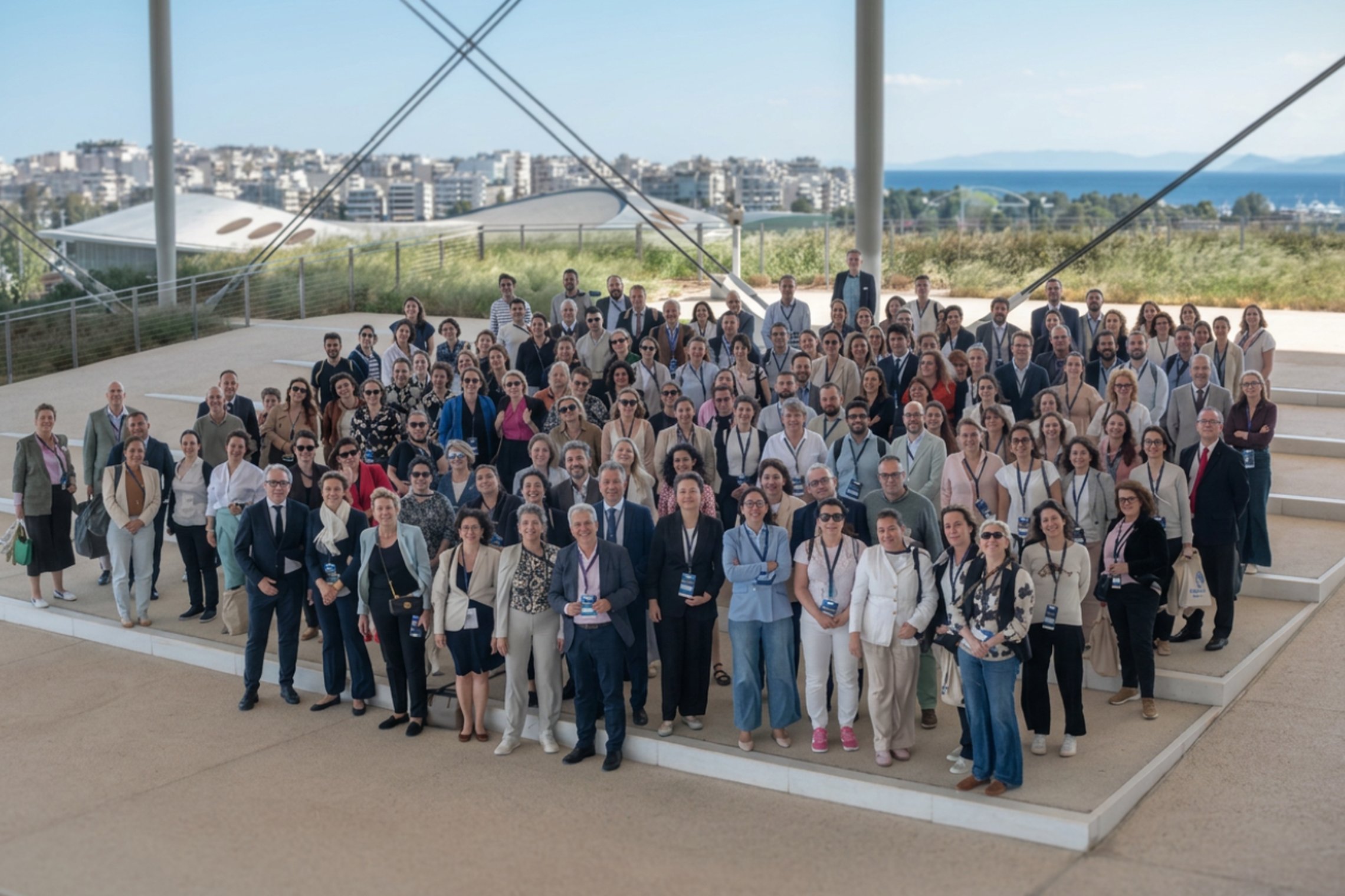 LT: Konferencijos dalyvių bendra nuotrauka lauke po metaline stoginės konstrukcija, fone – jūra ir miestas.  EN: Group photo of conference attendees outdoors under a metal canopy, with the sea and city in the background. 