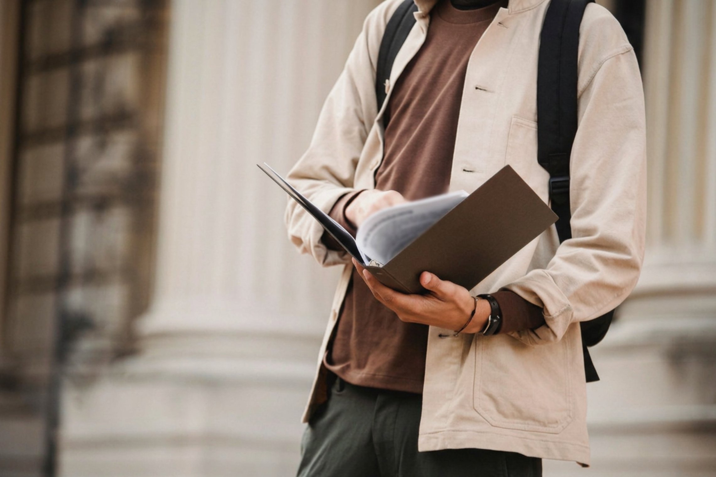 Lietuviškai: Studentas su kuprine laiko atvirą aplanką prie universiteto pastato. English: Student with a backpack holding an open folder outside a university building